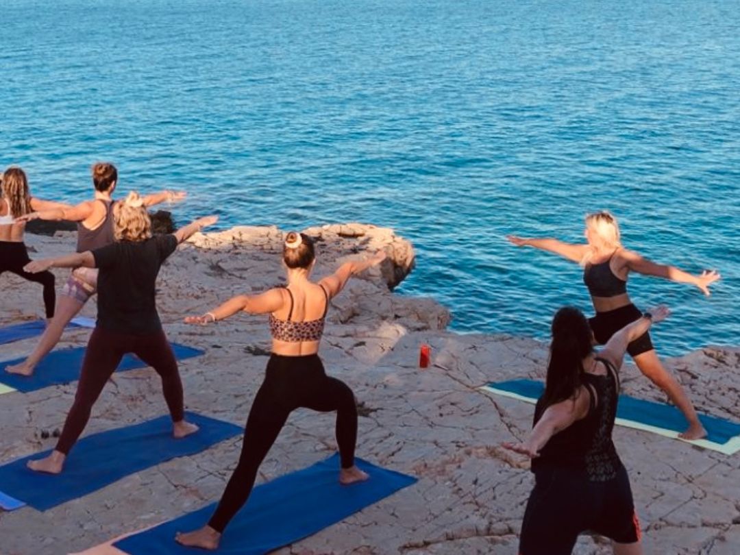 Women practicing Warrior 2 yoga pose on the beach by the blue sea